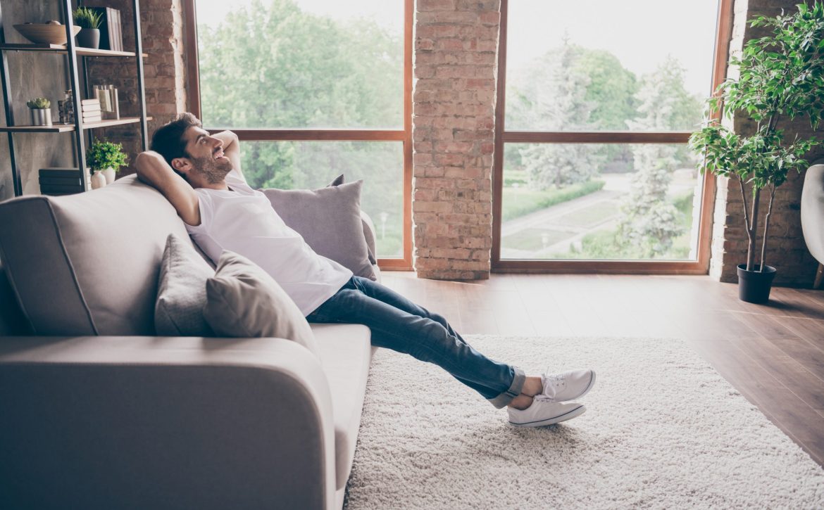 Profile photo of mixed race guy sitting cozy sofa holding hands behind head relaxing homey weekend mood looking dreamy new repair wear casual outfit flat loft living room indoors